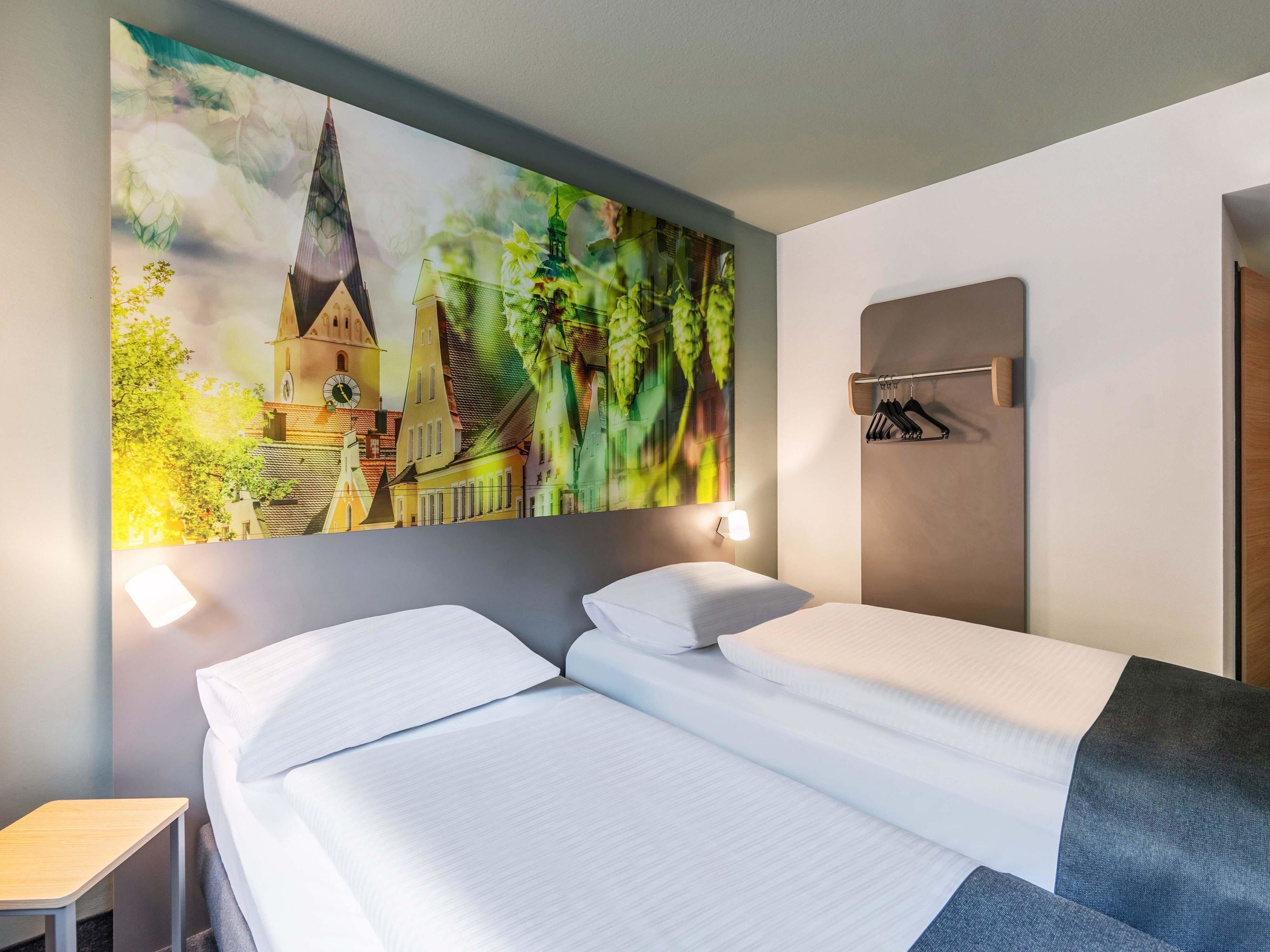 A hotel room with a bed, a nightstand, a wall-mounted coat rack, and a large cityscape painting.