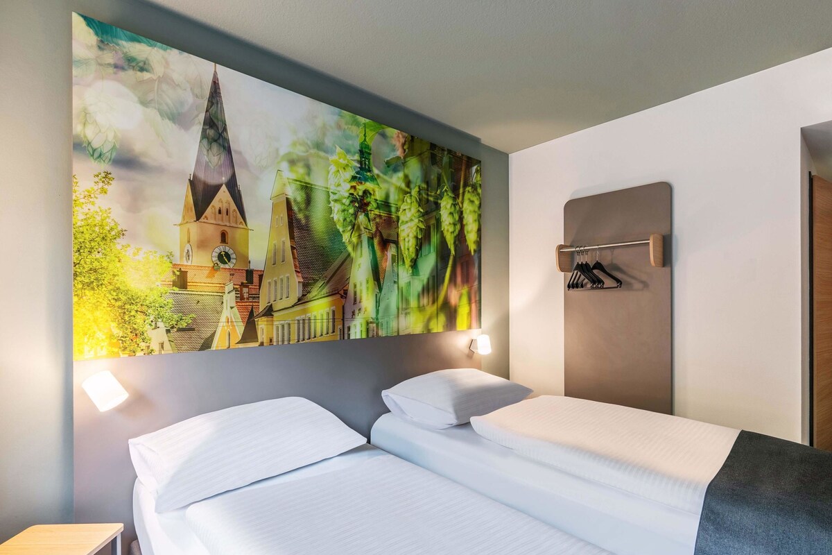 A hotel room with a bed, a nightstand, a wall-mounted coat rack, and a large cityscape painting.