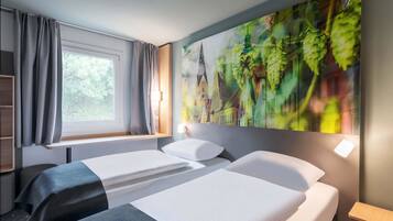 A hotel room with two beds, a window with curtains, a wall-mounted lamp, and a large cityscape painting.