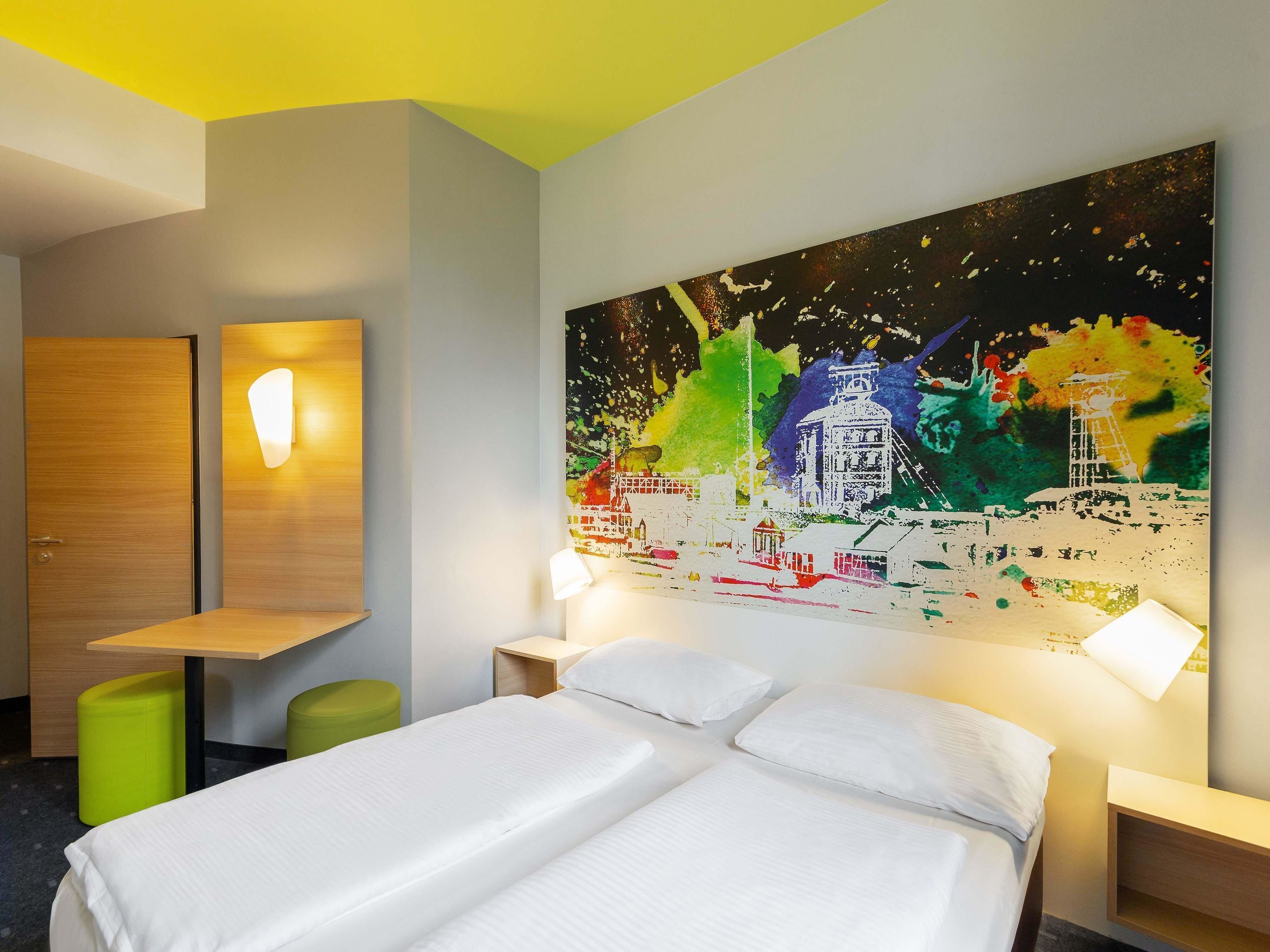 Ein Hotelzimmer mit einem Bett, zwei Nachttischen, einem grünen Hocker, einem Holztisch und einem großen, abstrakten Gemälde an der Wand.