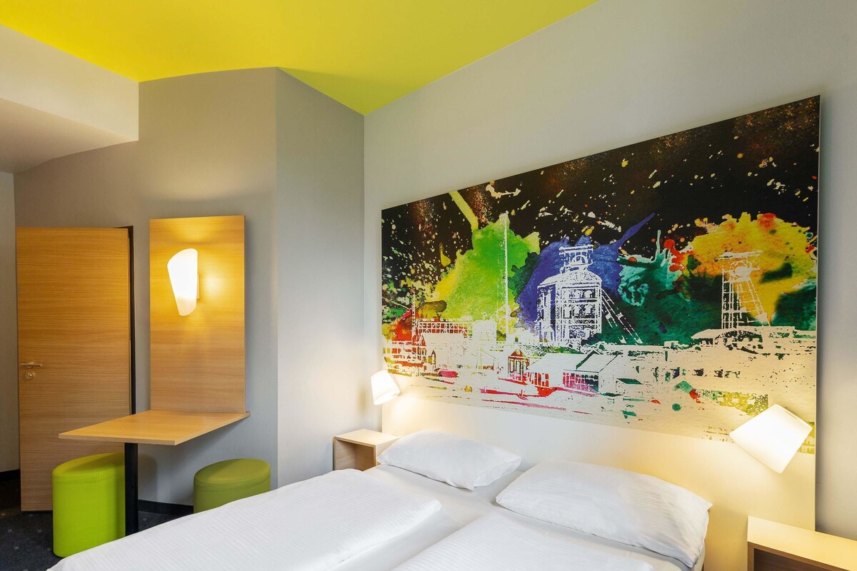 A hotel room with a bed, two bedside tables, a green stool, a wooden desk, and a large abstract painting on the wall.