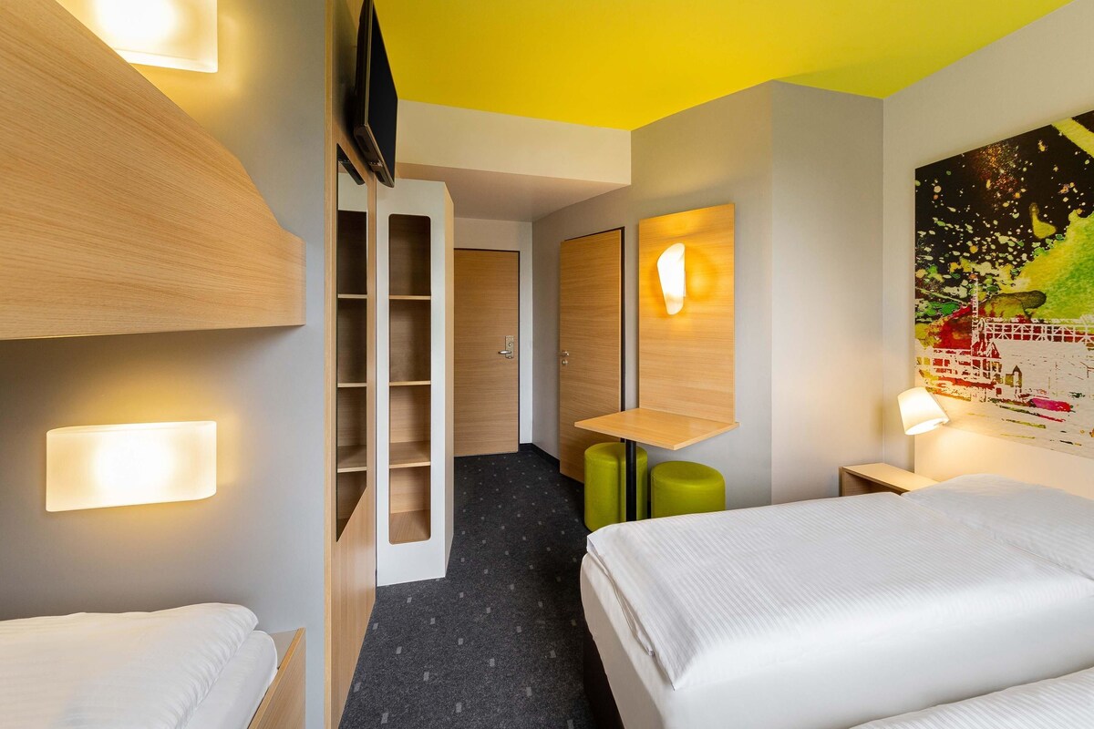 A hotel room with a bunk bed, a desk with a chair, a small table, a wall-mounted TV, and a painting on the wall.