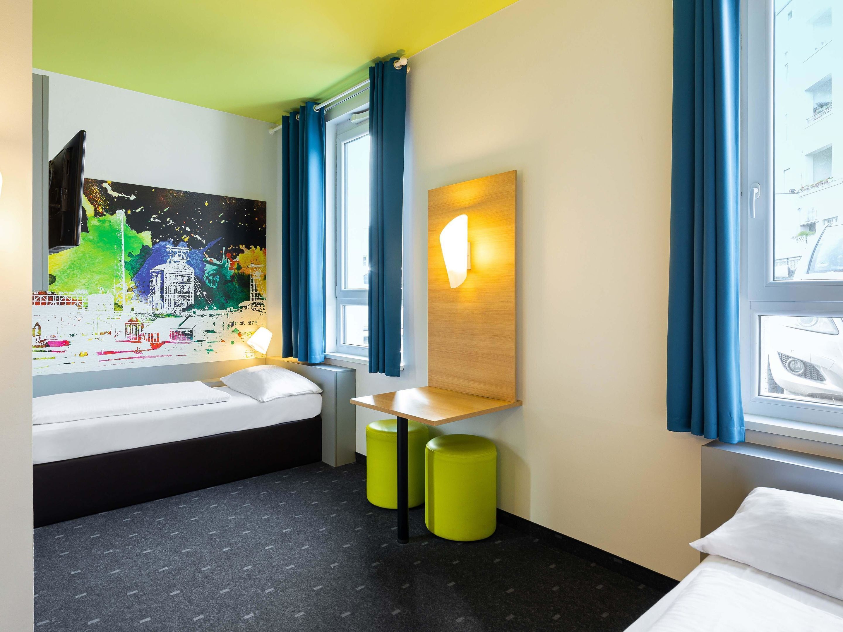 A modern hotel room with a bed, a desk, a colorful mural, and large windows with blue curtains.