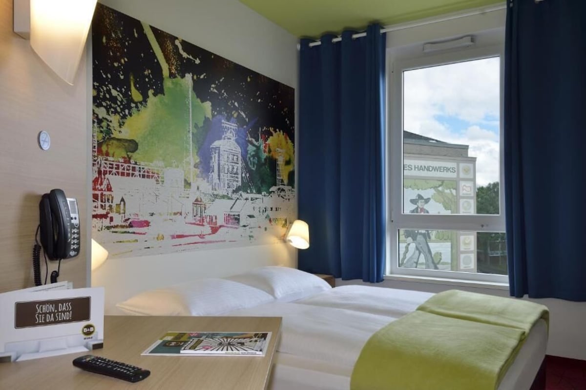 Une chambre d’hôtel comprenant un lit, un bureau avec un téléphone et une télécommande, une lampe fixée au mur et une fenêtre avec des rideaux.