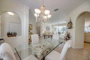 A dining area with a glass table, white chairs, a chandelier, and a view of the kitchen. - 16684 Wellington Pet Friendly Pool Home by Beach (Fort Myers)