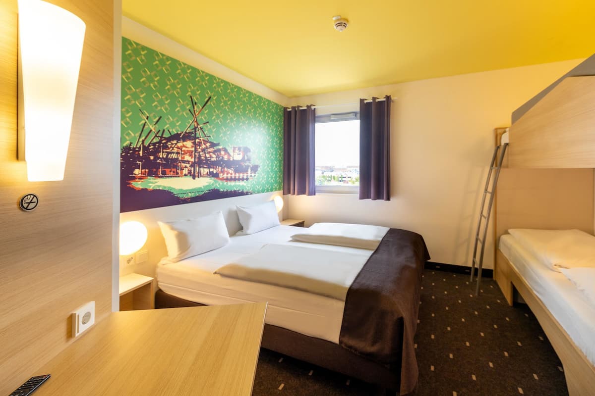 Una habitació d'hotel amb un llit, una taula de treball, una làmpada, una finestra amb cortines i un mural a la paret.