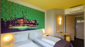 Une chambre d’hôtel avec deux lits, un tableau au mur, une table de chevet, une petite table, une lampe fixée au mur et une porte.