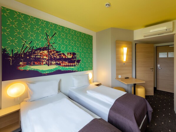 Habitación de hotel con dos camas, un cuadro en la pared, una mesita de noche y una lámpara de pared, además de una puerta.