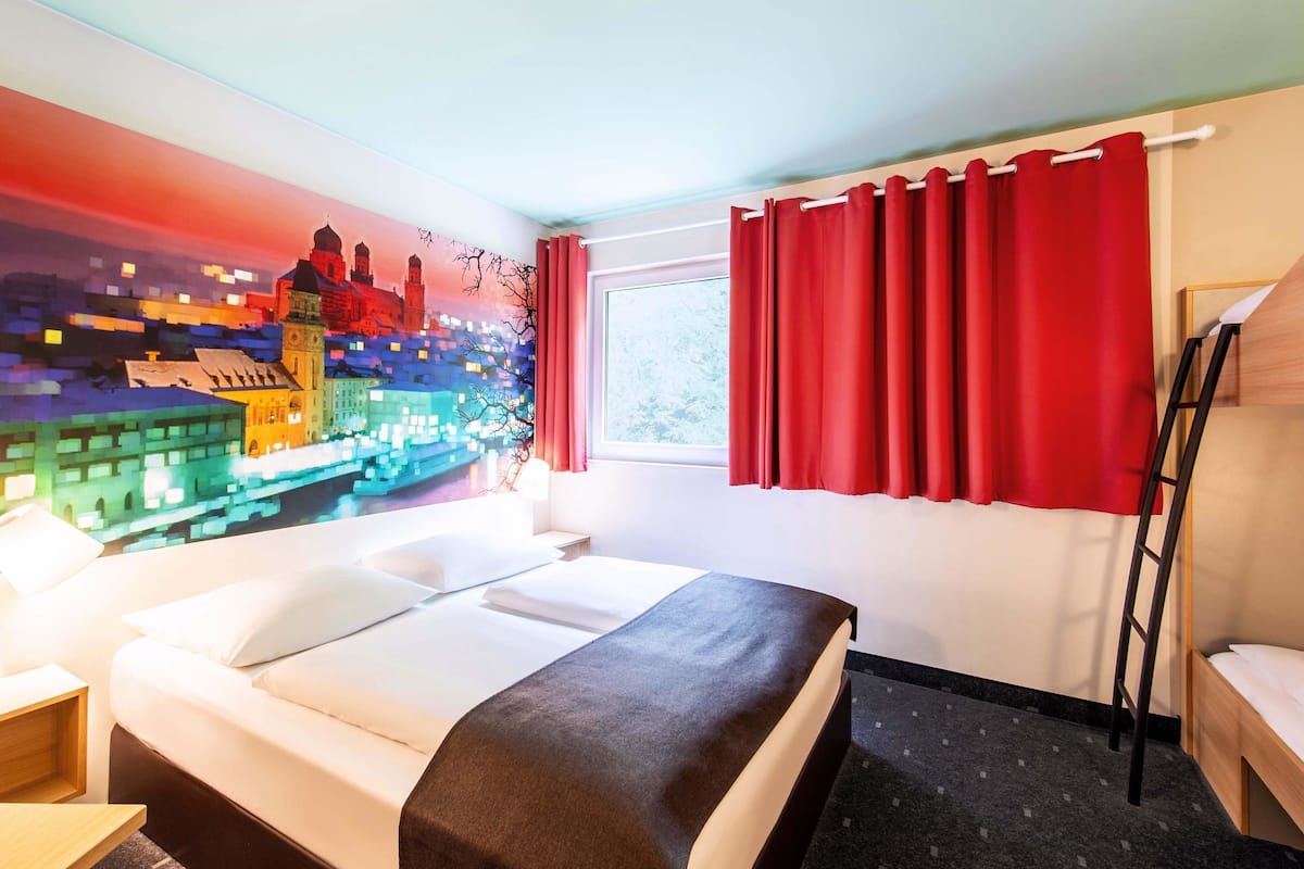 Hotellihuone, jossa on sänky, yöpöytä, työpöytä, punainen verho ja ikkuna, josta on näkymä rakennuksiin.