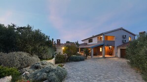 Une maison moderne à deux étages, dotée d’une allée pavée et entourée de jardins aménagés et d’arbres matures.