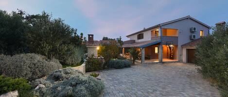A modern two-story house with a paved driveway, surrounded by landscaped gardens and mature trees.