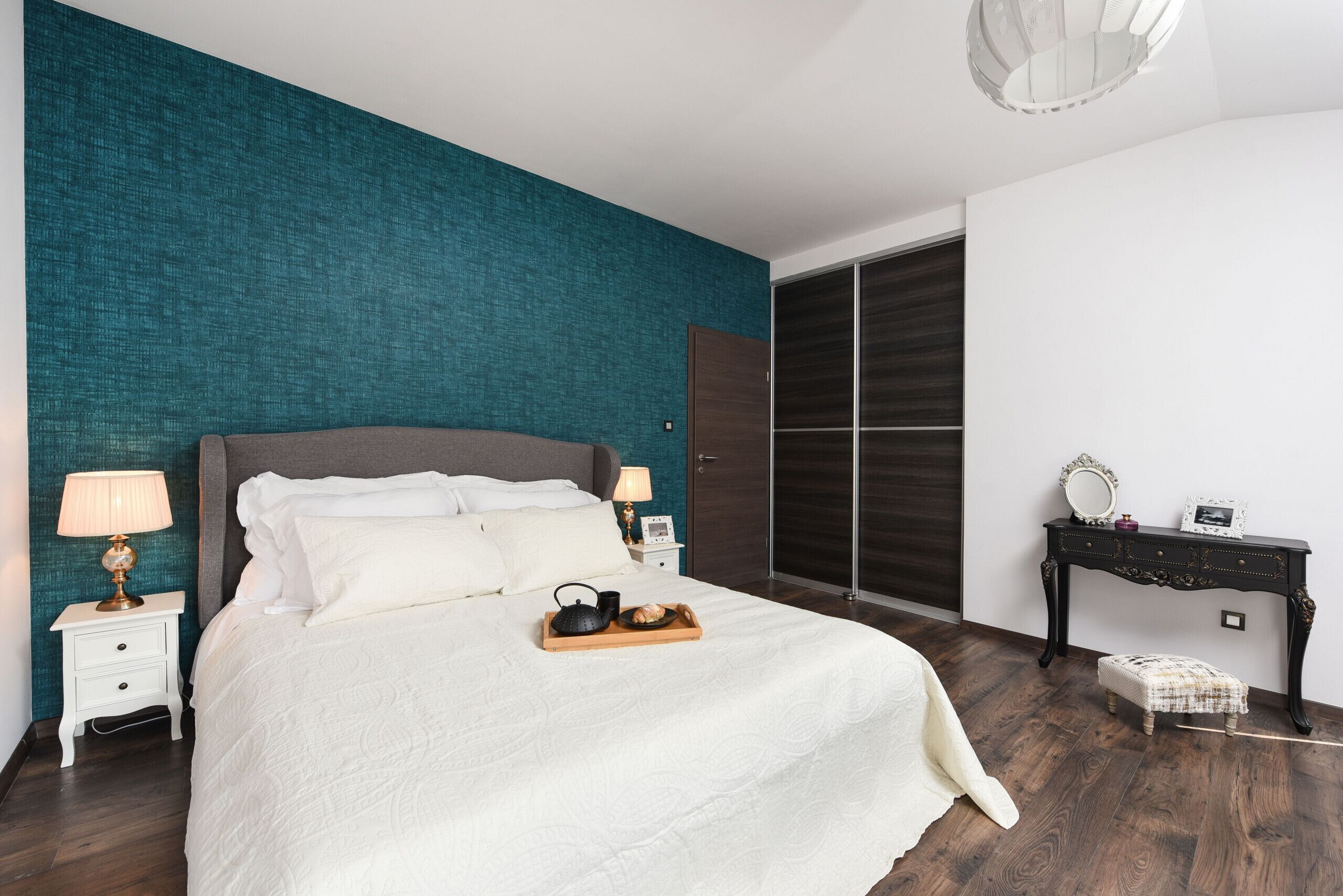 A neatly made bed with pillows and a tray of items, a nightstand with a lamp, a dark wooden wardrobe, and a small table with a mirror and picture frame.