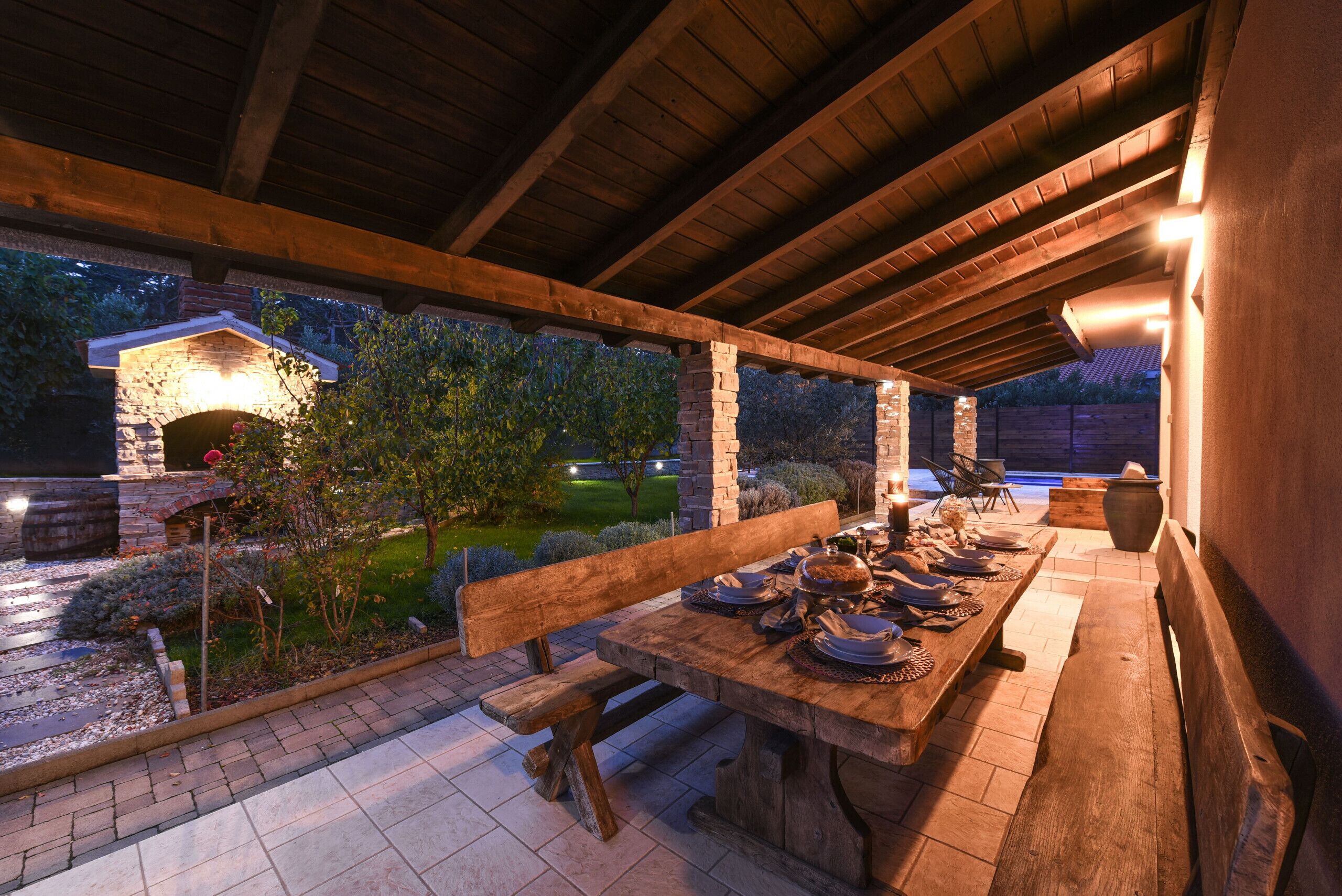 Un área de comedor al aire libre con chimenea de piedra, bancos de madera y una larga mesa preparada para una comida.