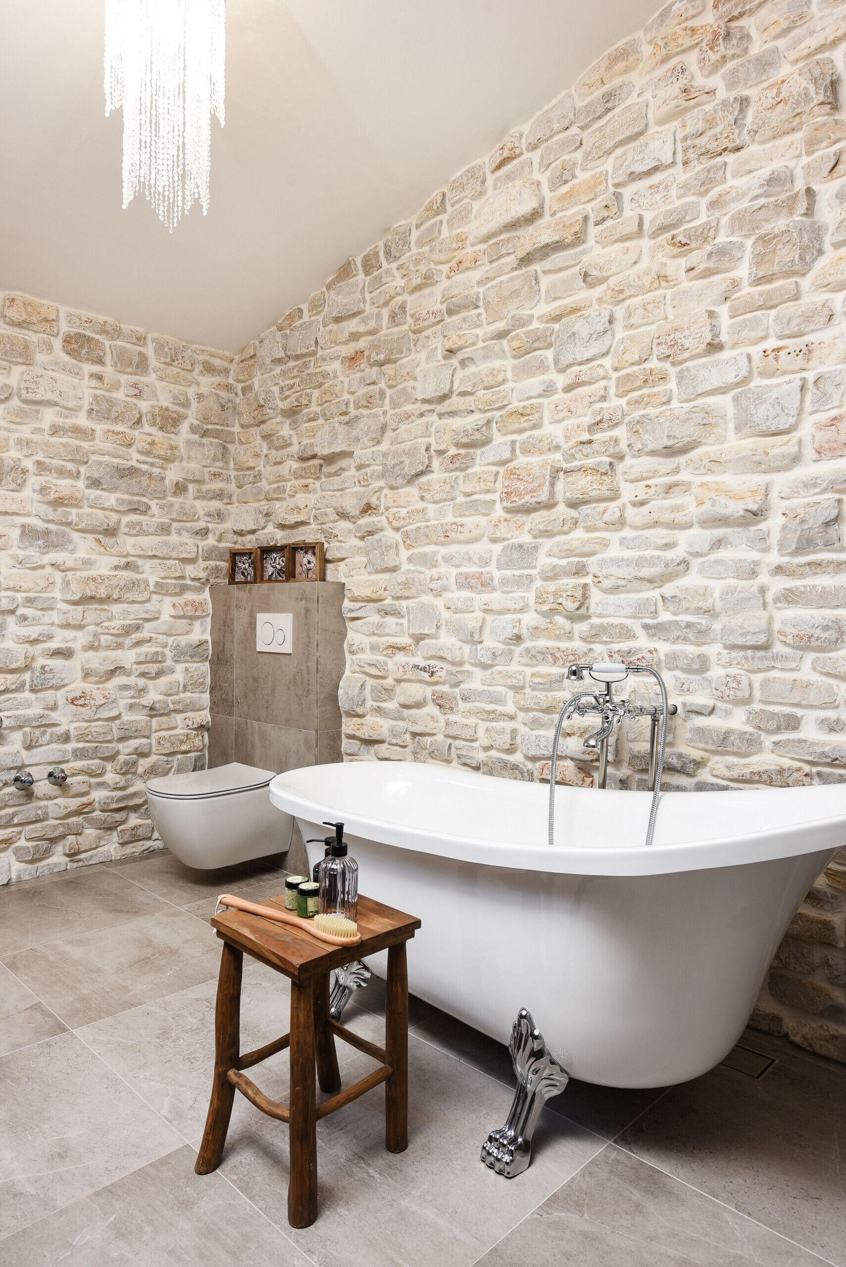 A bathroom with a stone wall, a freestanding bathtub, a wooden stool, and a toilet.