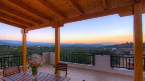 Un balcone in legno con un tavolo e sedie, affacciato su un paesaggio suggestivo con montagne ed edifici.
