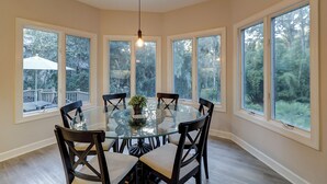 Un comedor con una mesa de vidrio rodeada de sillas, ventanas amplias y una lámpara colgante.