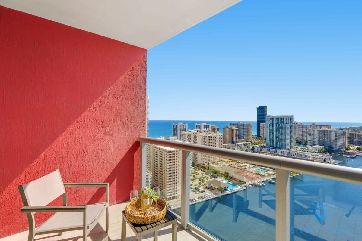 A balcony with a red wall, a chair, and a table with drinks. A cityscape and water are visible in the background.
