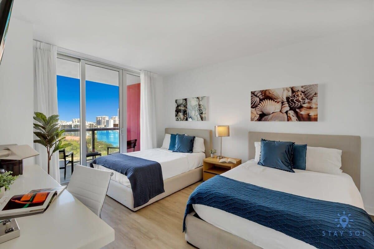 A hotel room with two beds, a desk, and a view of the city.