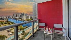 A balcony with a red wall, two chairs, a table with a bottle and glass, and a view of a city skyline and water body. - Studio w/Infinite Views & Pool  - Near Beach (Hallandale Beach)