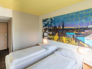 Een hotelkamer met twee enkele bedden, een groot stadsgezichtschilderij aan de muur en een deur links.