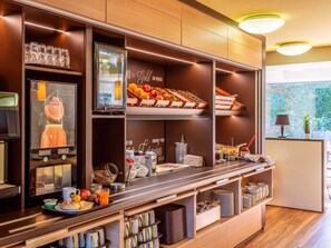 A breakfast bar with a variety of food items, a coffee machine, and a window with a view of greenery. - B&B Hotel Aachen-Würselen (Wuerselen)