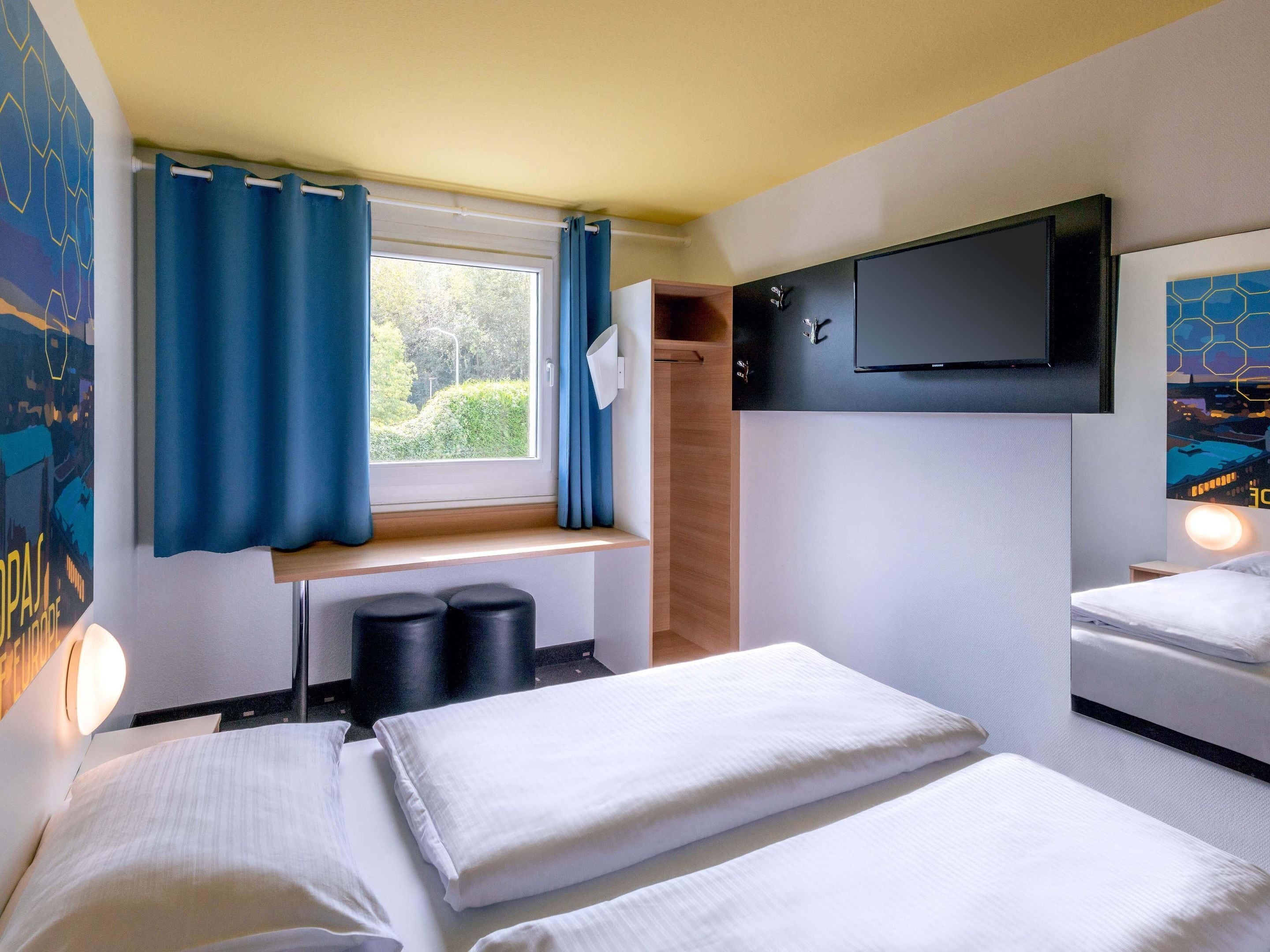 A hotel room with a bed, a window with curtains, a TV mounted on the wall, and a small seating area with stools.