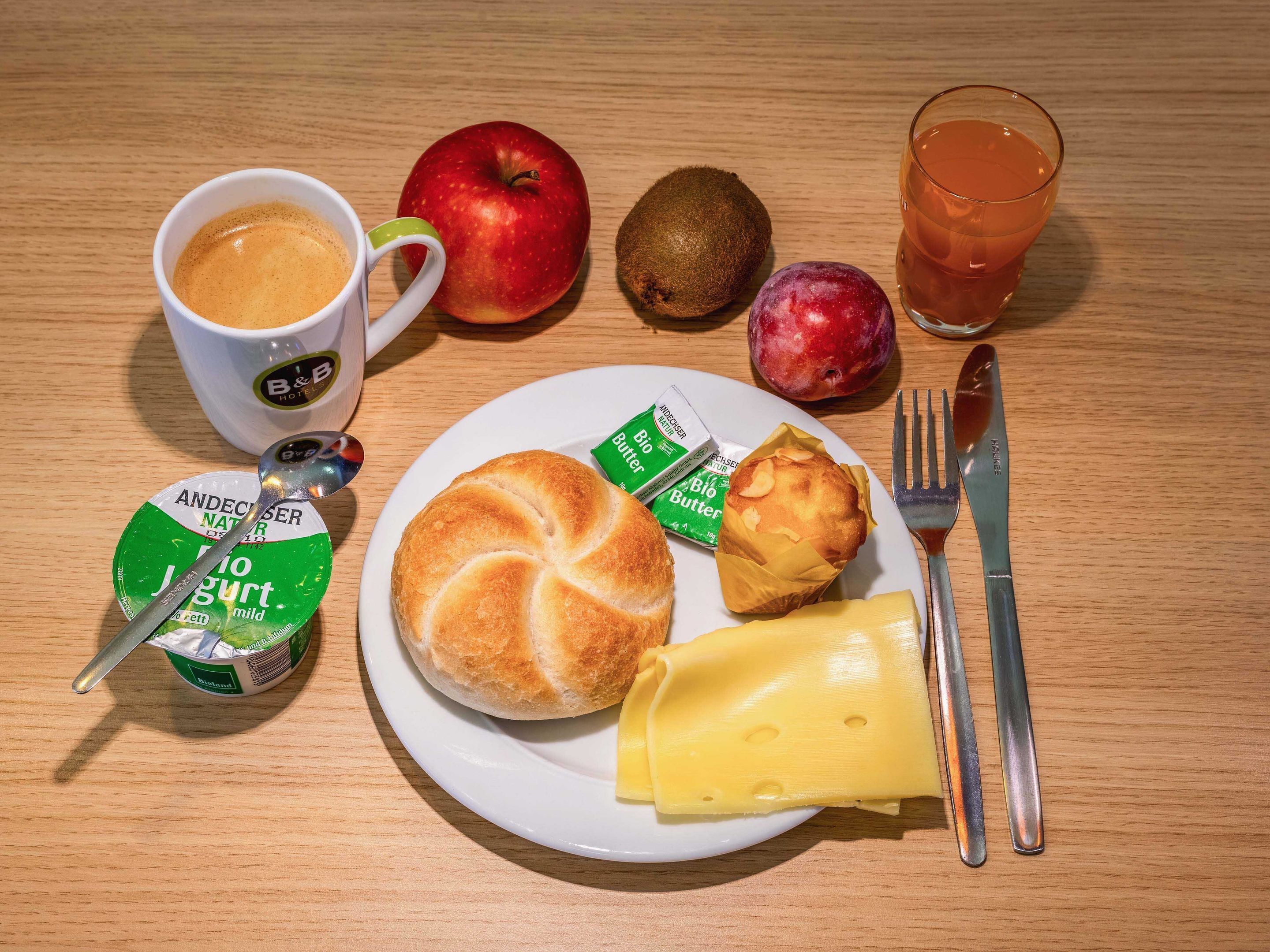 Un plato de desayuno con un croissant, queso, mantequilla, un muffin, una manzana, un kiwi, un vaso de jugo y una taza de café.