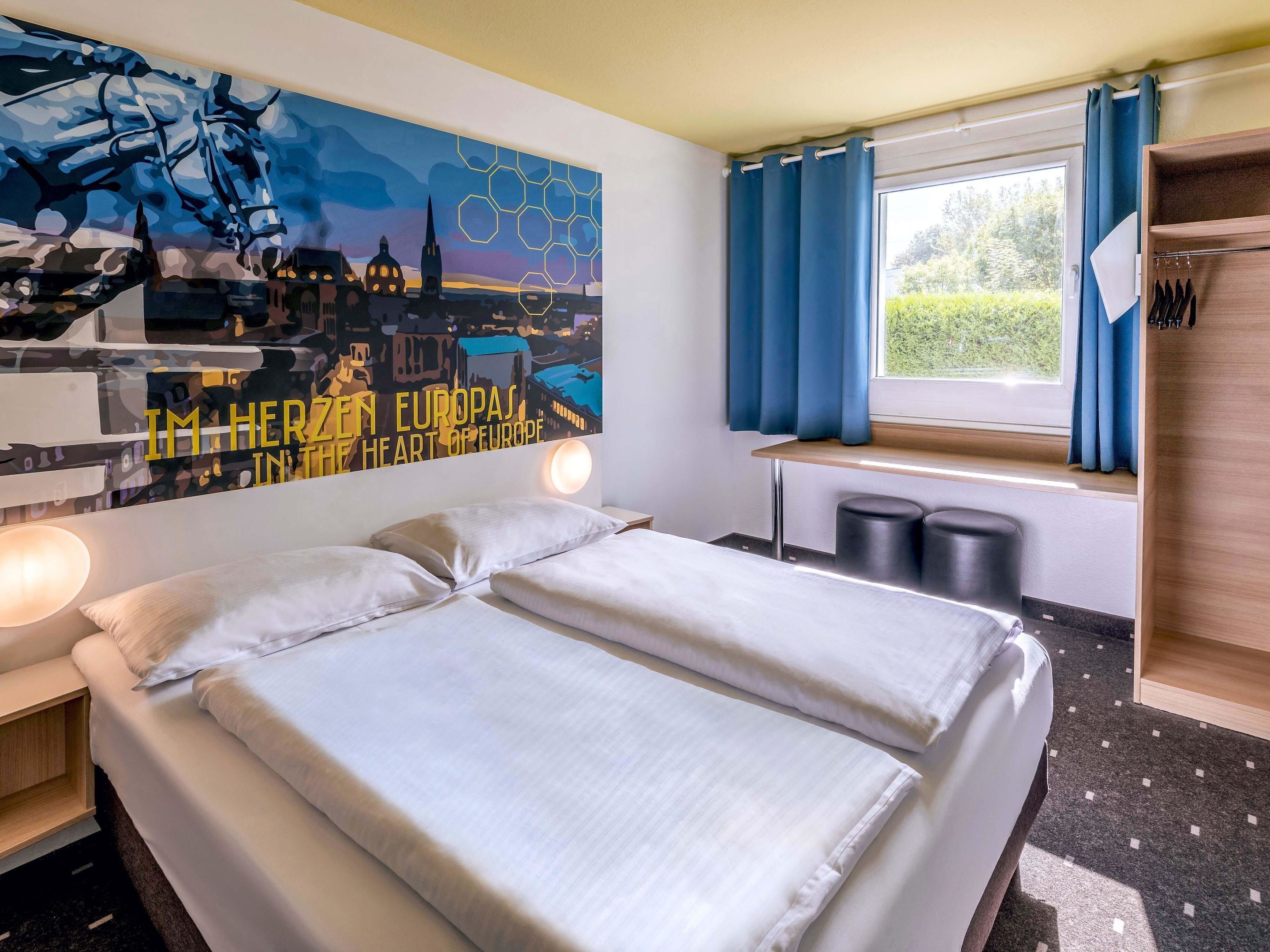 A hotel room with a bed, a nightstand, a wall art depicting a cityscape, a window with curtains, and a wooden shelf with items.