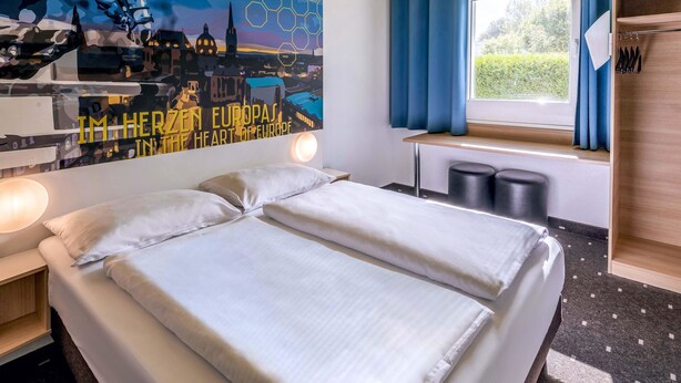 Une chambre d’hôtel comprenant un lit, une table de chevet, une œuvre d’art murale représentant un paysage urbain, une fenêtre avec des rideaux et une étagère en bois sur laquelle sont posés divers objets.