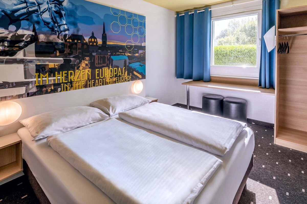 A hotel room with a bed, a nightstand, a wall art depicting a cityscape, a window with curtains, and a wooden shelf with items.