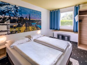 A hotel room with a bed, a nightstand, a wall art depicting a cityscape, a window with curtains, and a wooden shelf with items. - B&B Hotel Aachen-Würselen (Wuerselen)