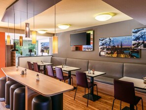 Un restaurant moderne avec un intérieur élégant, offrant différents types de sièges, notamment des banquettes et des tables, ainsi qu’un grand téléviseur fixé au mur.