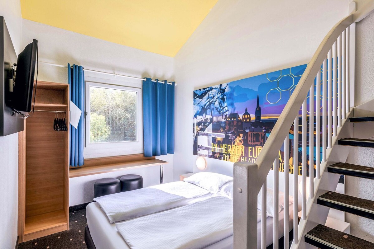 Une chambre d’hôtel avec un lit, un bureau, une fenêtre avec des rideaux, un tableau au mur et un escalier.
