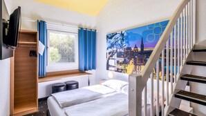 Une chambre d’hôtel avec un lit, un bureau, une fenêtre avec des rideaux, un tableau au mur et un escalier.