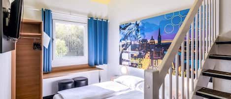 A hotel room with a bed, a desk, a window with curtains, a painting on the wall, and a staircase.