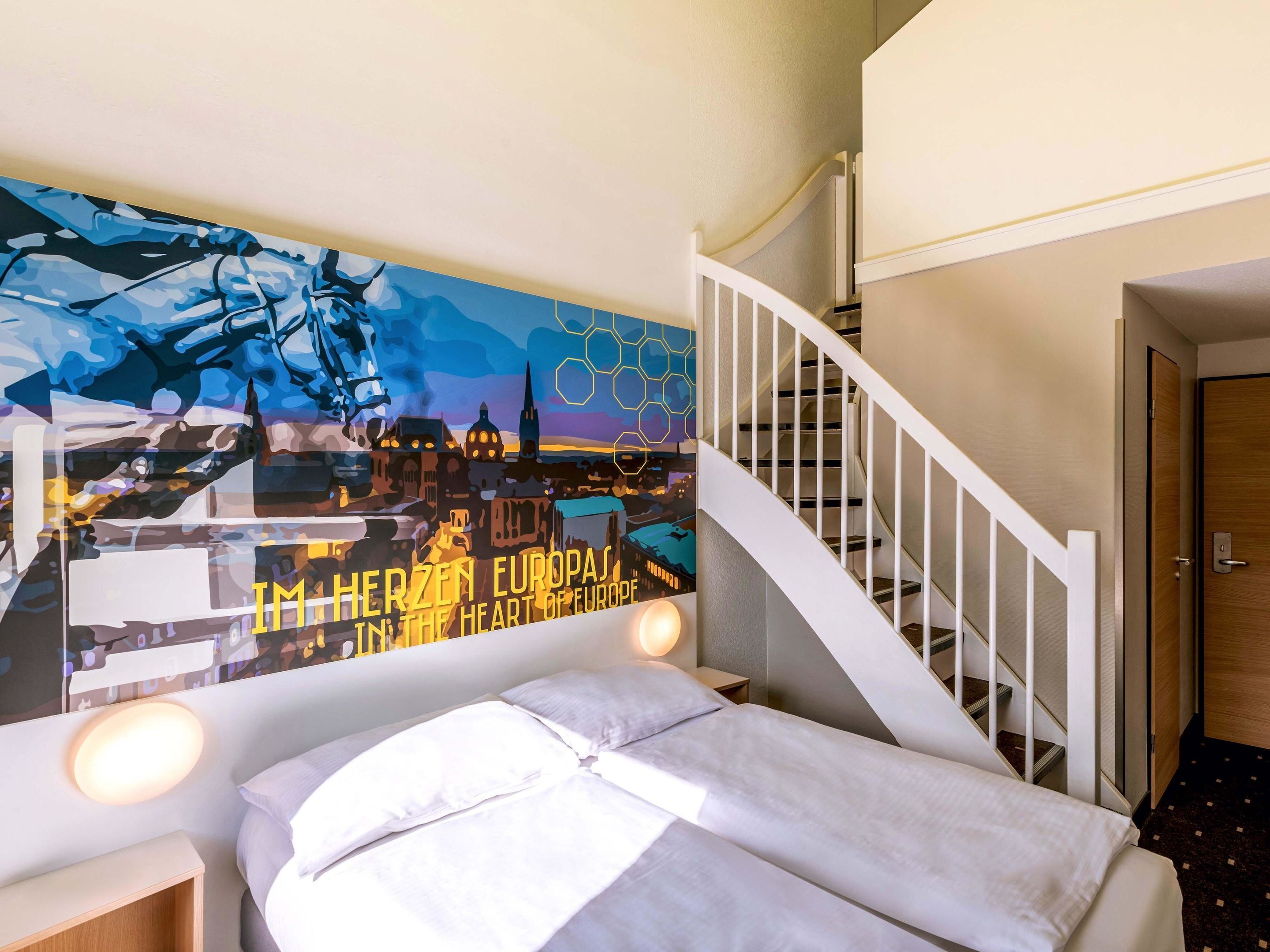 Een hotelkamer met een bed, een nachtkastje, een muur met een stadsgezicht schilderij en een trap.