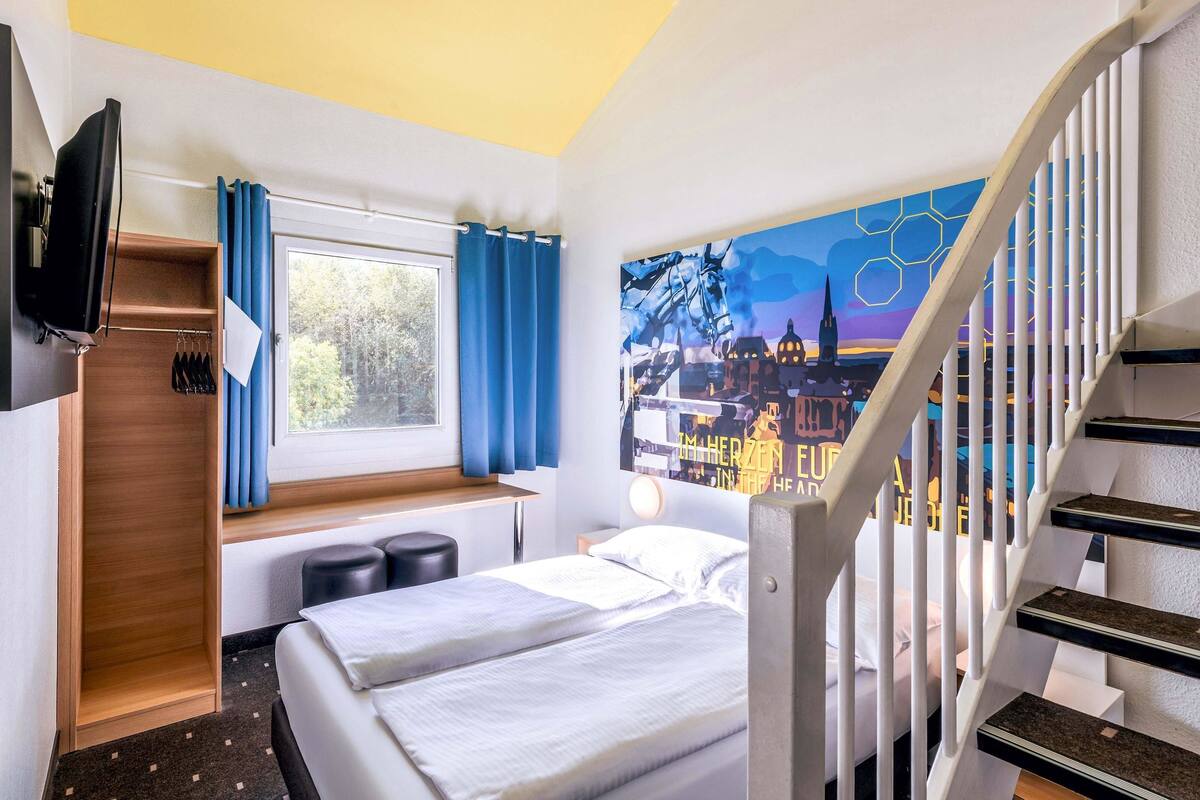 Une chambre d’hôtel avec un lit, un bureau, une fenêtre avec des rideaux, un tableau au mur et un escalier.