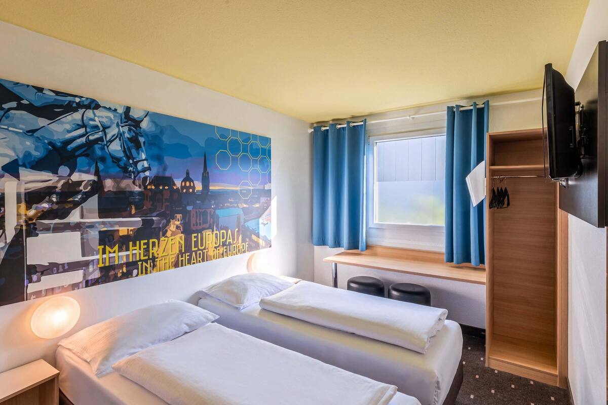 Une chambre d’hôtel avec deux lits, un bureau, une télévision et un grand tableau représentant un paysage urbain accroché au mur.