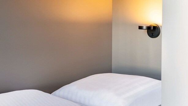 A hotel room with a bed, a wall-mounted light fixture, and a plain wall. - B&B Hotel Berlin-Airport (Schoenefeld)