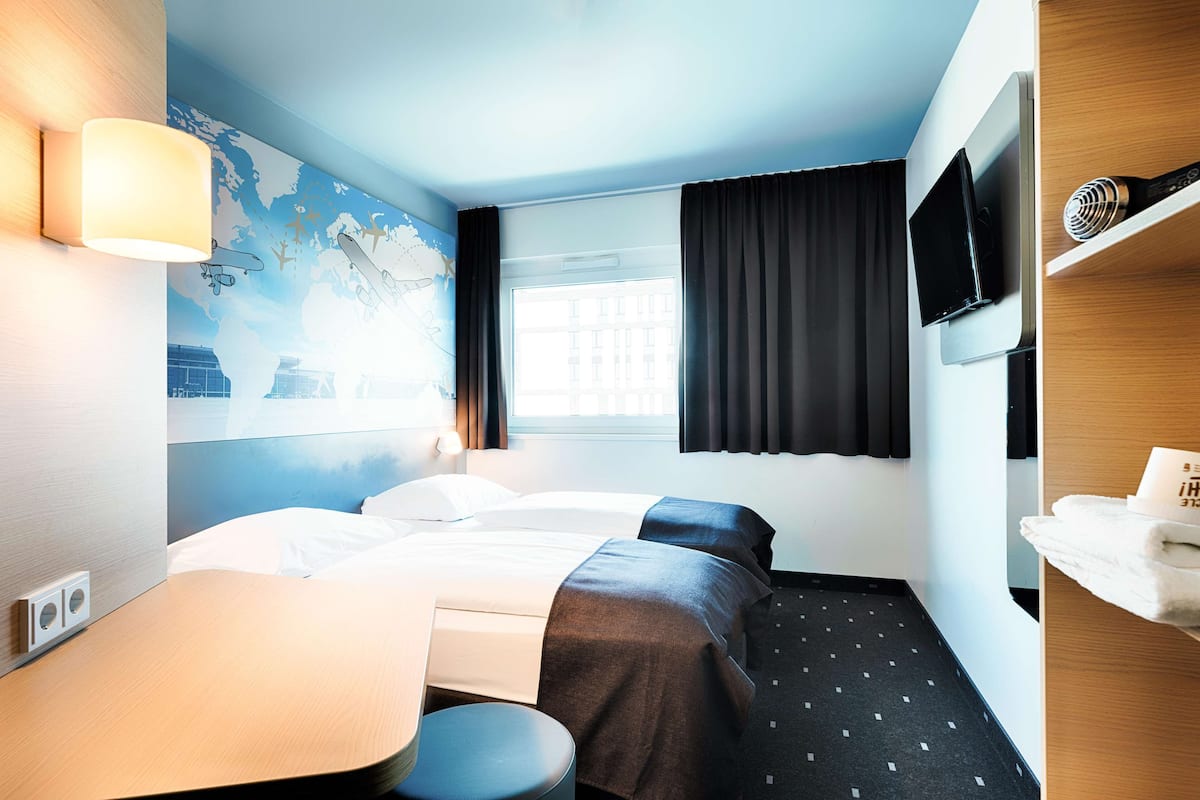 Habitación de hotel con dos camas, un escritorio, una silla, un televisor de pantalla plana, un mapa en la pared y una ventana con cortinas.