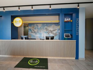 Un comptoir d’accueil d’un hôtel de la chaîne B&B, avec une fresque représentant une carte du monde, un espace d’accueil et un panneau indiquant la présence d’un bar.