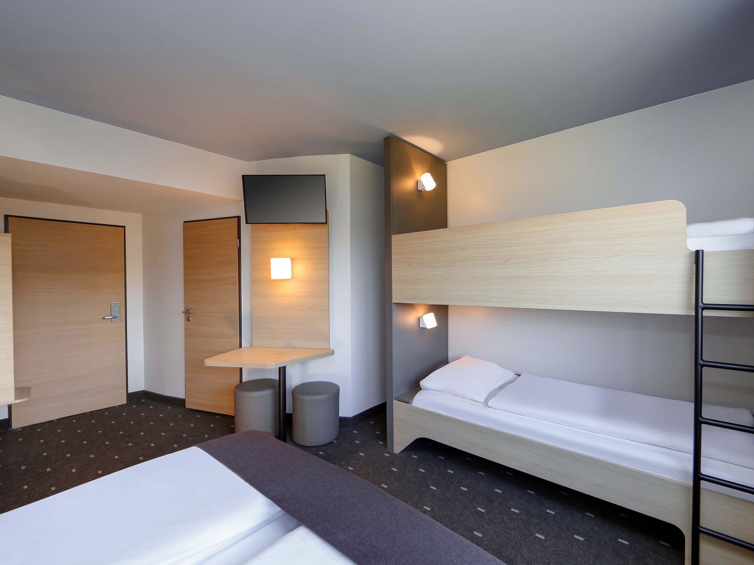 A hotel room with a bunk bed, a desk with a chair, and a small table. There are two doors, one of which is open.