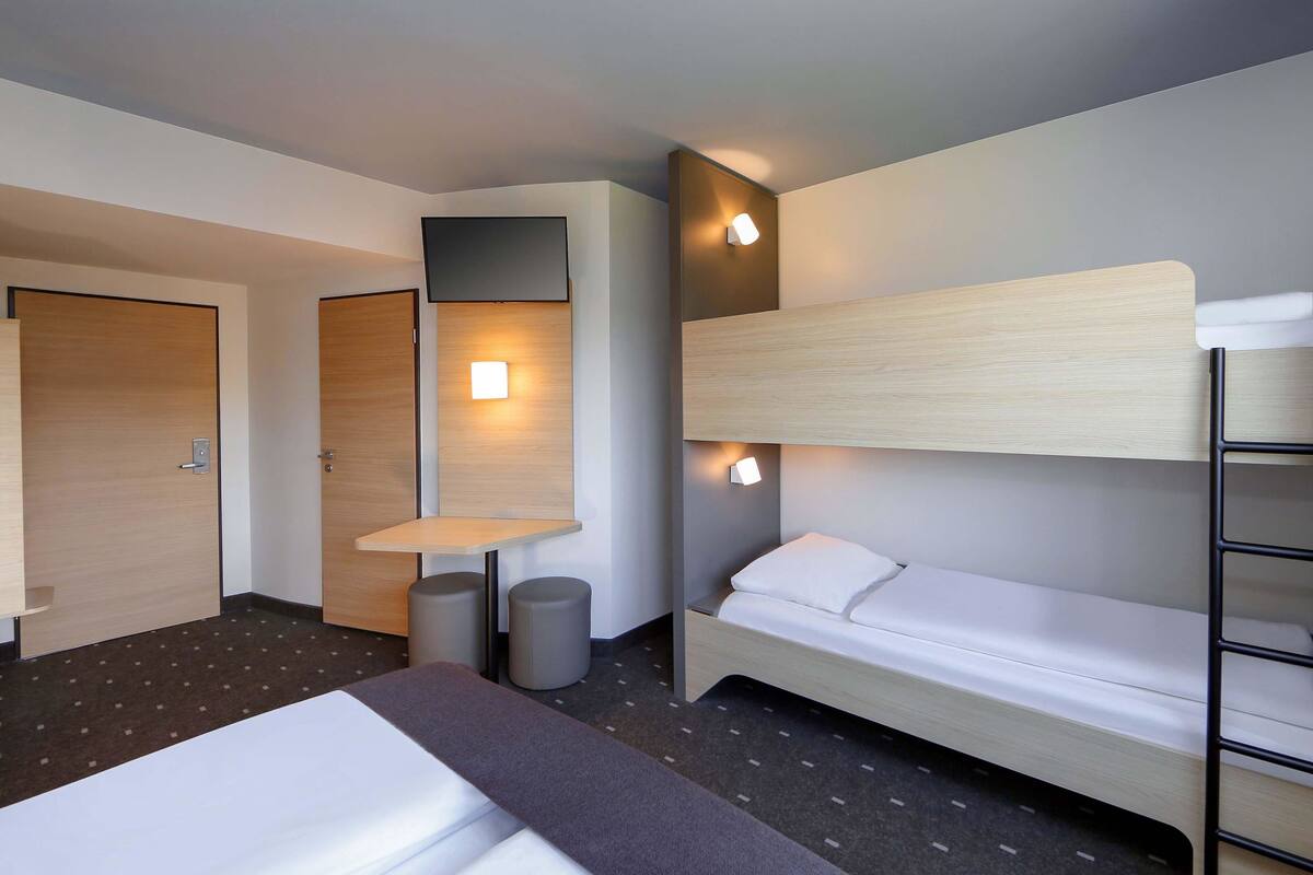 Habitación de hotel con una cama litera, un escritorio con silla y una mesita pequeña. Hay dos puertas, una de las cuales está abierta.