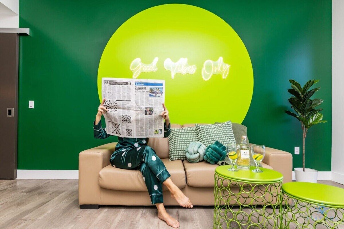 A person sitting on a sofa with a newspaper, a neon sign in the background, and a small table with drinks.