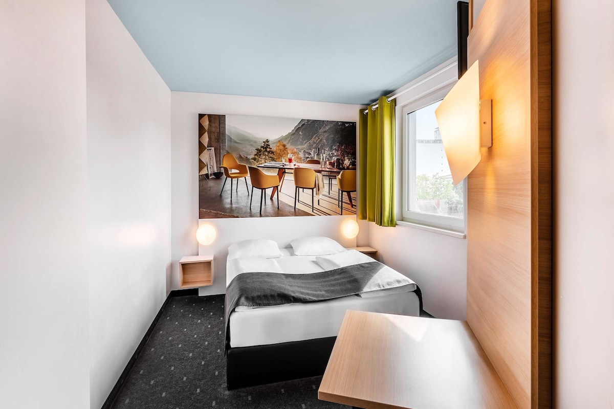 Una camera d'albergo moderna con un letto, un comodino e vista su un'area ristorazione attraverso un'ampia finestra.