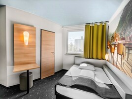 Una habitación de hotel moderna con una cama, una mesita de noche, una lámpara de pared, una ventana con cortinas y vistas a edificios.