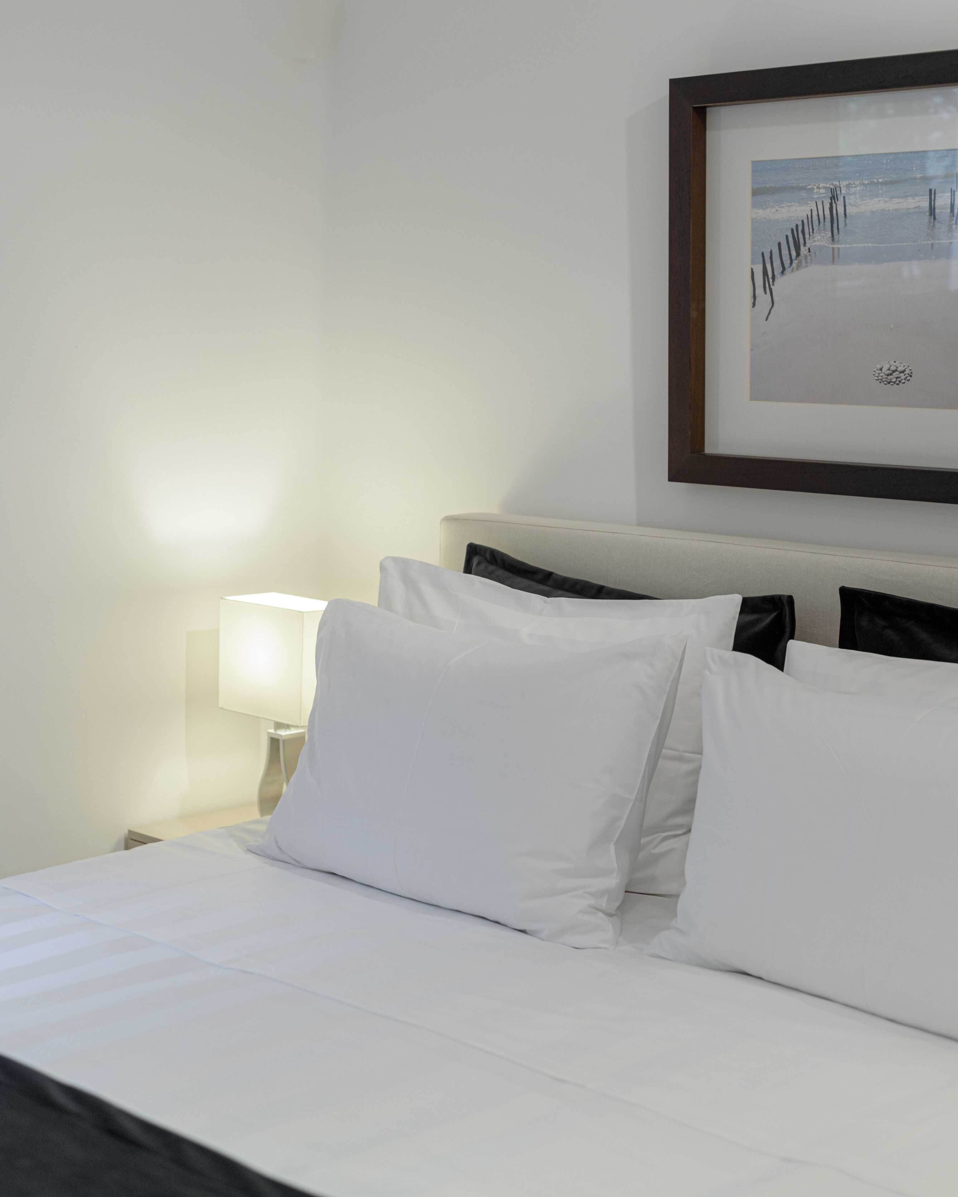 A hotel room with a neatly made bed, a framed picture on the wall, and a bedside lamp.