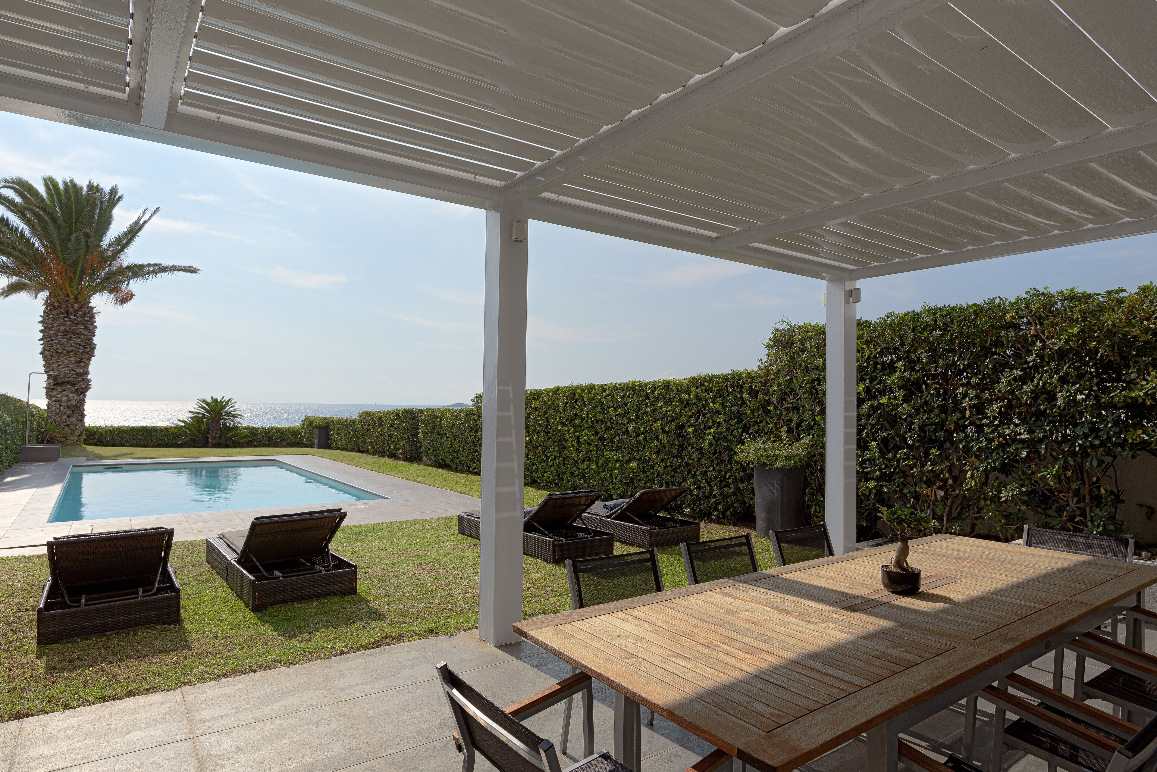 An outdoor patio with a pool, lounge chairs, a wooden table, and a view of the sea.