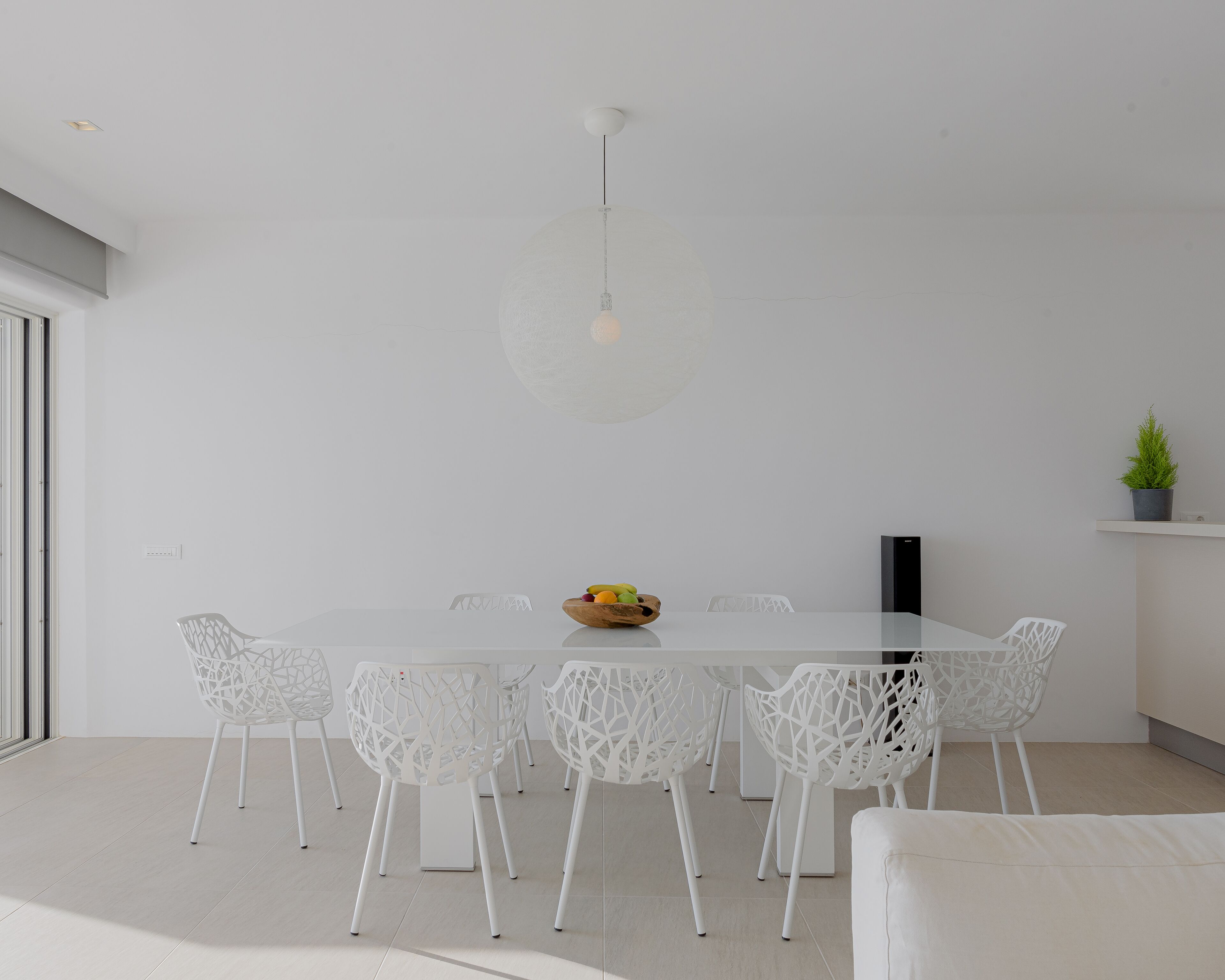 Et moderne spiseområde med et glasbord, hvide stole, en hængende lampe og en potteplante.