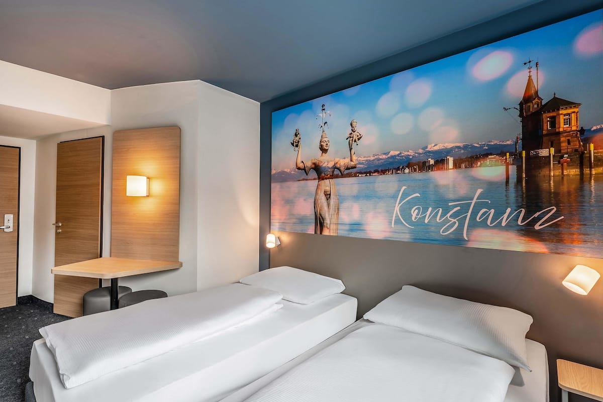 A hotel room with two beds, a wall-mounted TV displaying a scenic view, and a small table with a lamp.